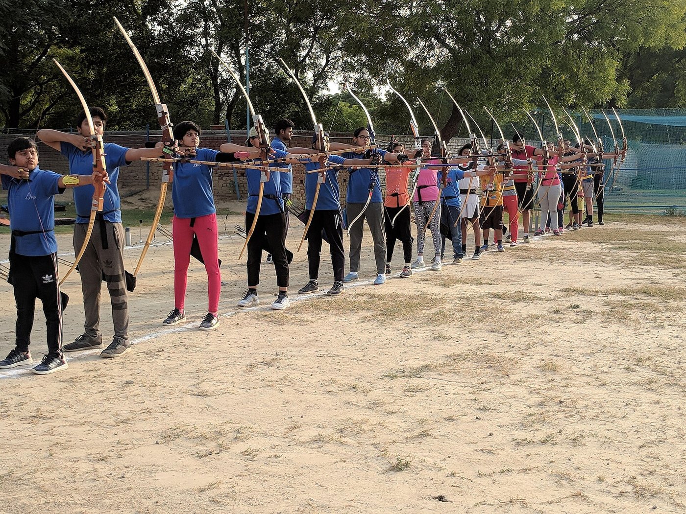 A long line of young archers drawing recurve bows at an outdoor range in India