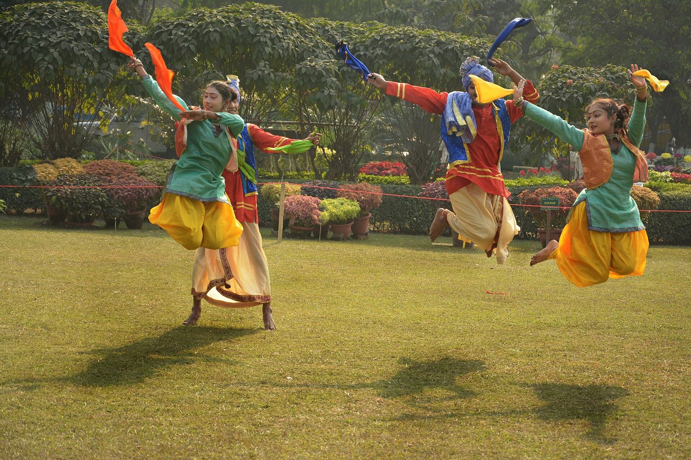 Three bhangra dancers in bright green, orange and yellow mid-leap on a lawn, scarves flying