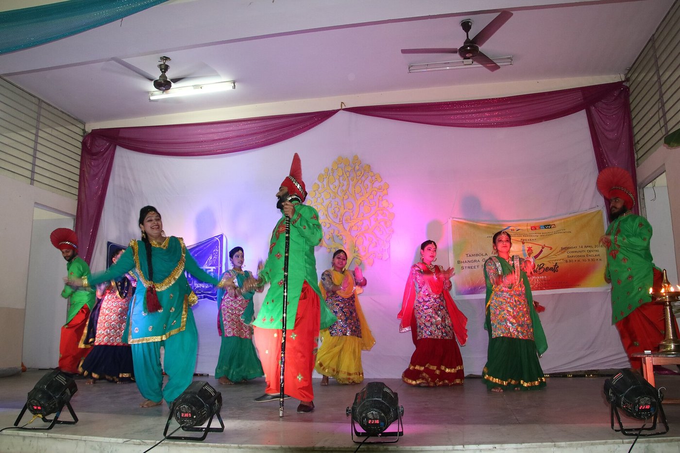 Performers in Punjabi dress, turbans and bright suits, dancing on an auditorium stage under coloured lights