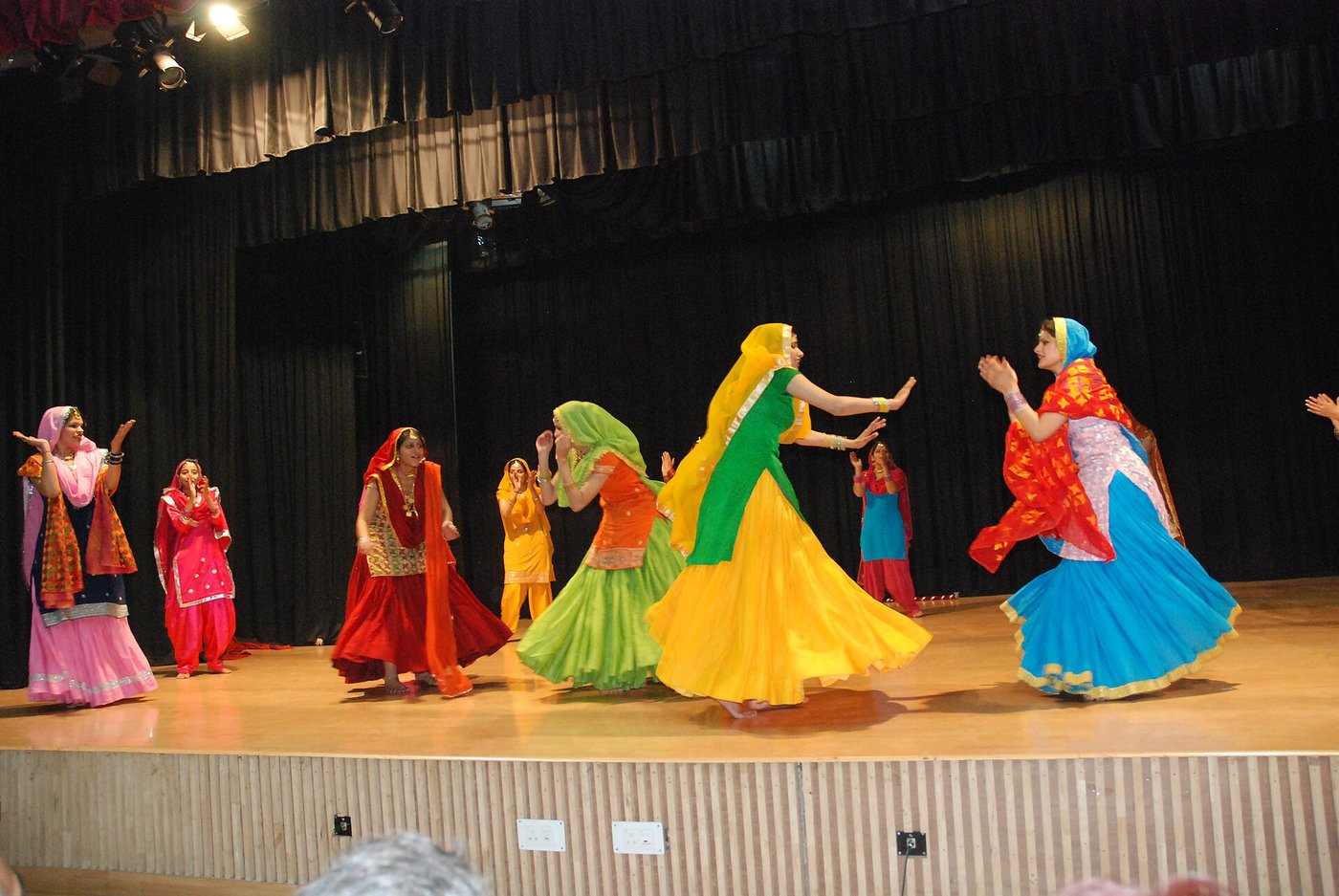 Women performers in green, yellow, orange and blue Punjabi suits and dupattas dancing giddha on a stage