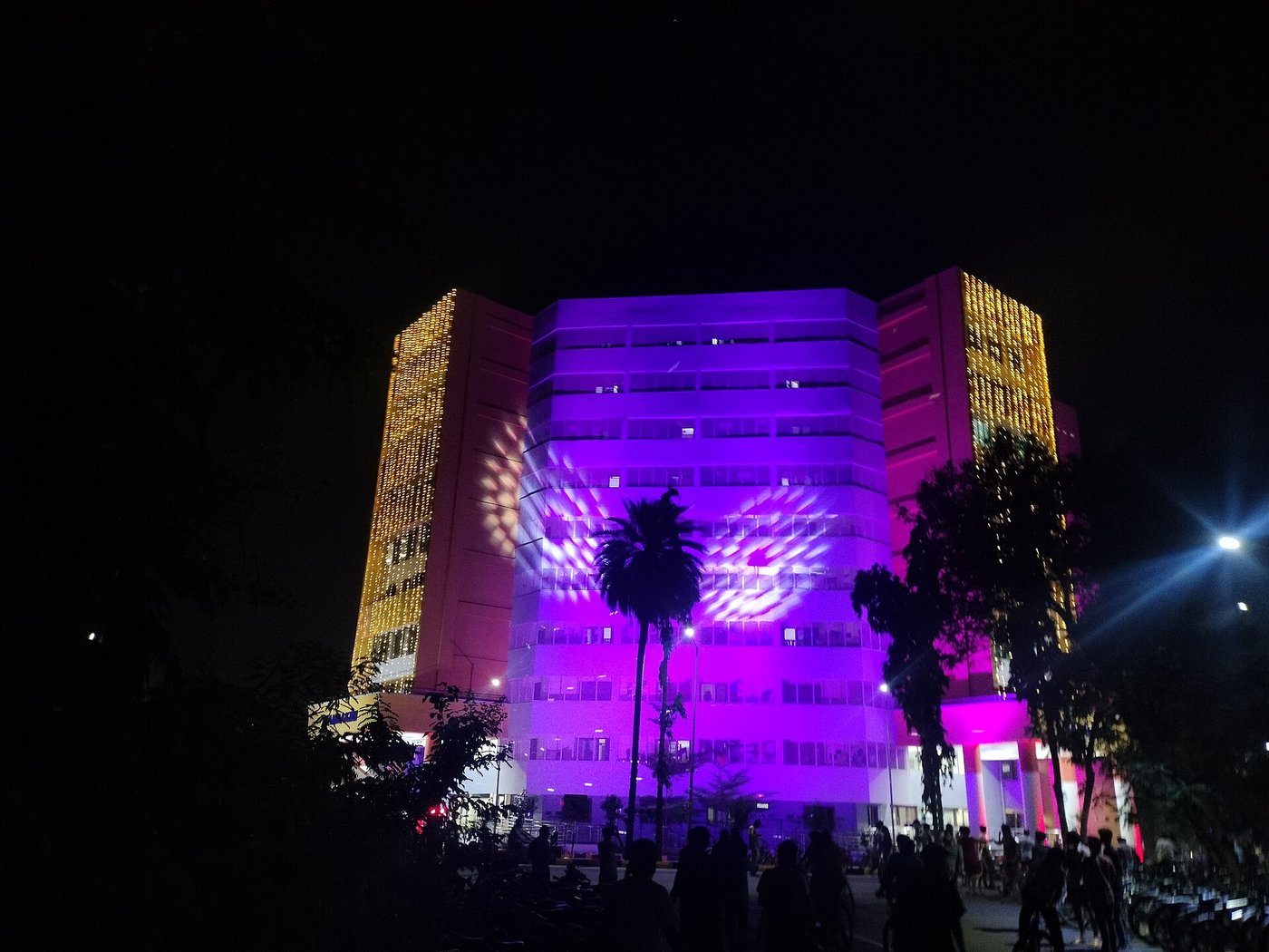 A hostel tower at IIT Madras lit up in purple and gold at night with students gathered below