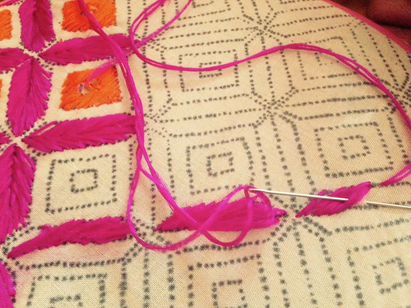 Close-up of hot pink and orange phulkari thread being stitched into a geometric pattern on cream cloth